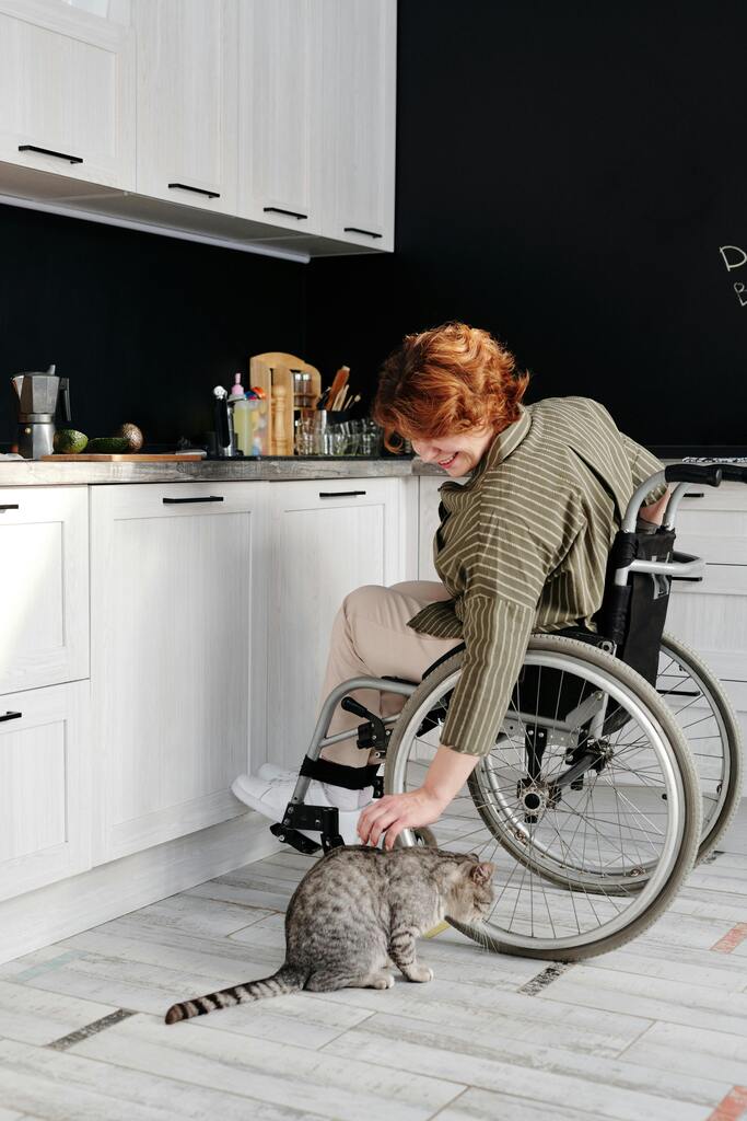 Handicap et logement : Une personne en situation de handicap sourit en caressant un chat tabby, installée confortablement dans une cuisine moderne aménagée pour l’accessibilité.